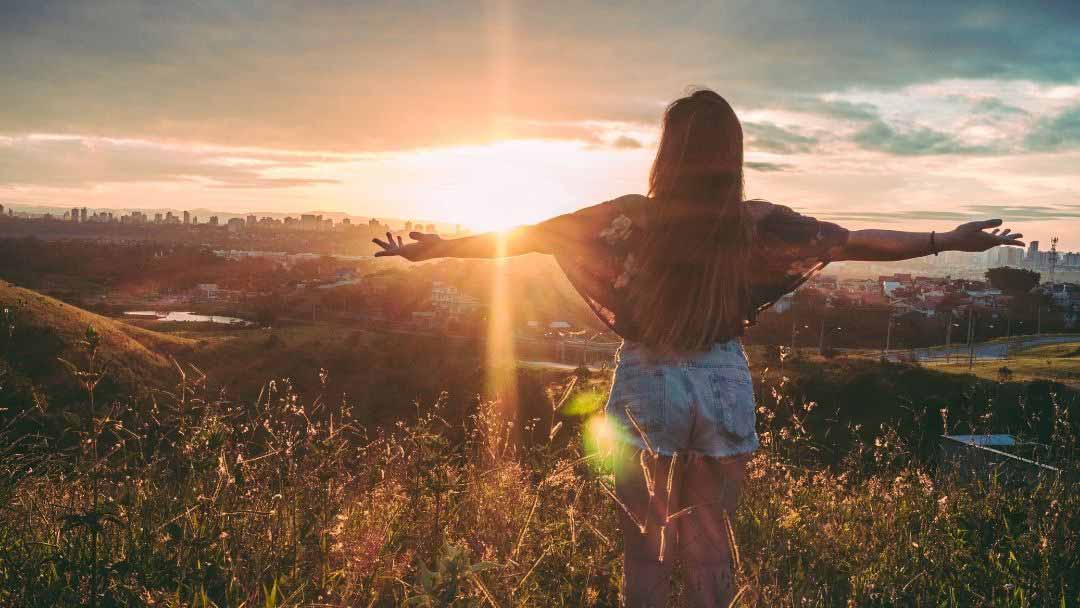 Nuori nainen seisoo kukkaniityllä auringon laskiessa selin kameraan, kädet levällään.