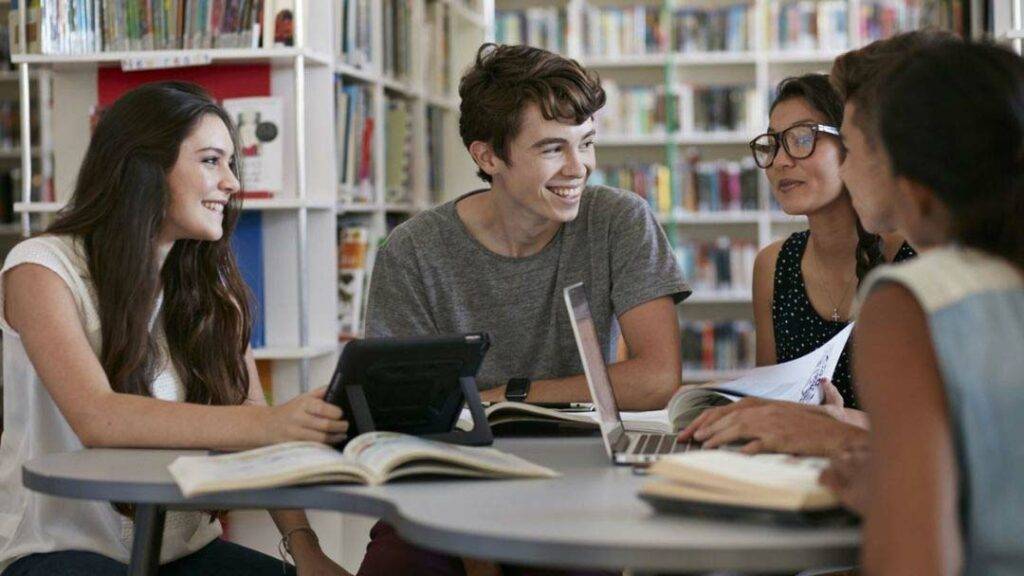 Kolme nuorta naista istuu nuoren miehen kanssa hymyillen kirjastossa päydän ääressä ja tekevät läksyjä