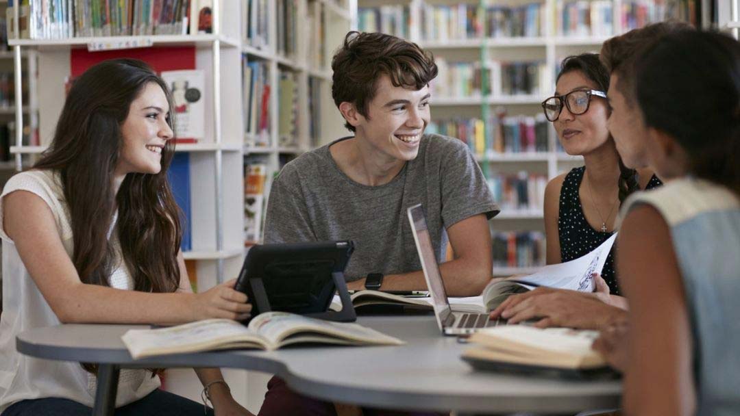 Kolme nuorta naista istuu nuoren miehen kanssa hymyillen kirjastossa päydän ääressä ja tekevät läksyjä