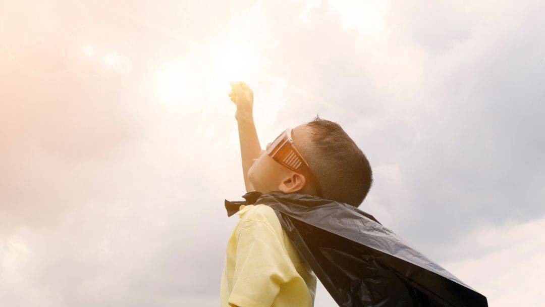 Pikkupoika leikkii supersankaria viitta päällä ja laskettelulasit silmillä, käsi kohti taivasta yrittäen ottaa auringosta kiinni