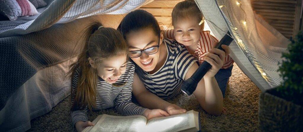 Äiti lukee peittomajassa kahdelle lapselle satuja taskulampun valossa.